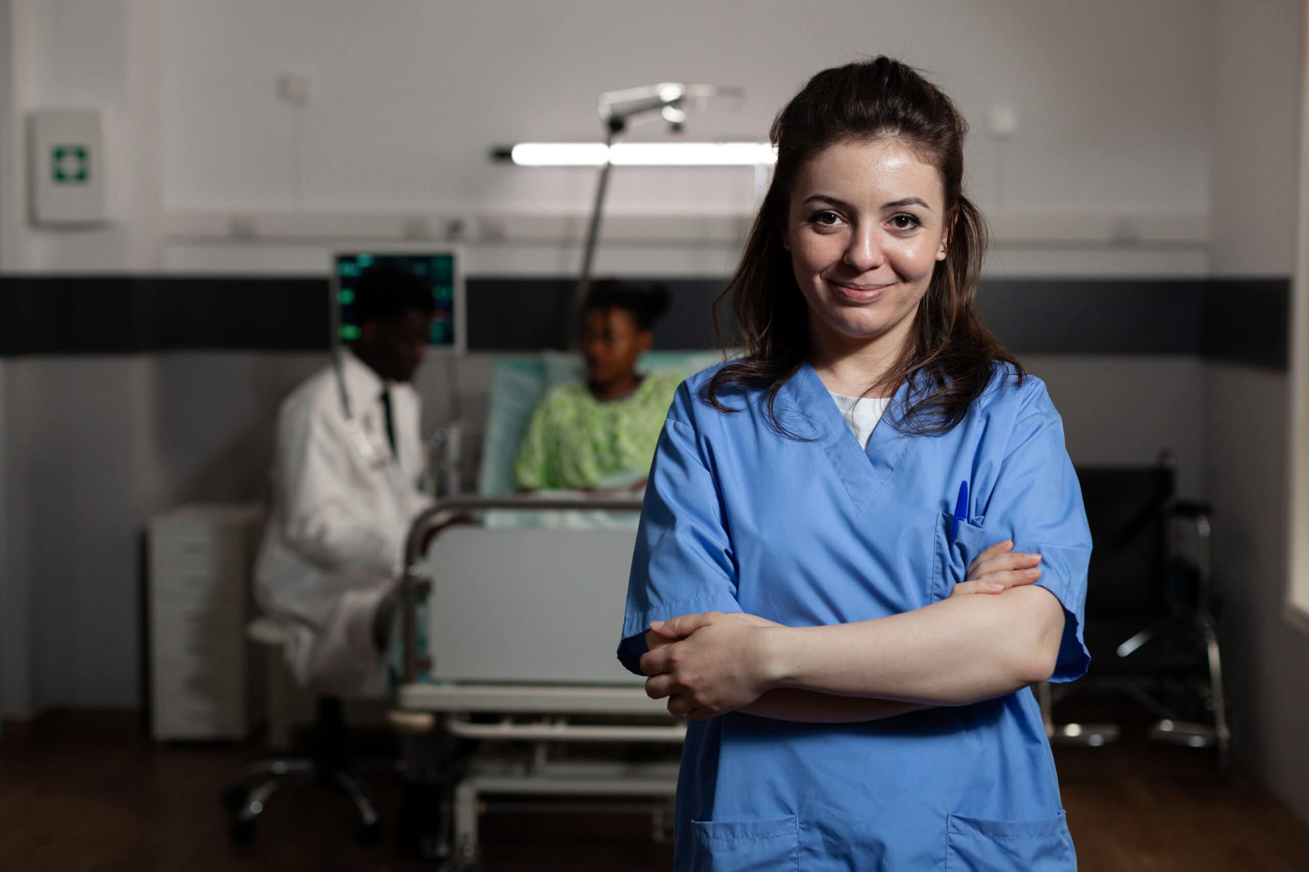 Healthcare professional in scrubs smiling in a hospital room with a patient in the background.